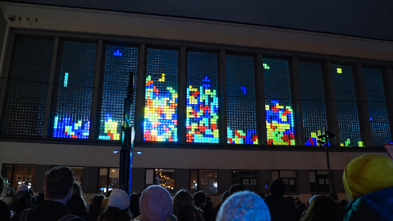 A crowd in winter hats watching falling-blocks games glow in the school’s glass-block windows at night