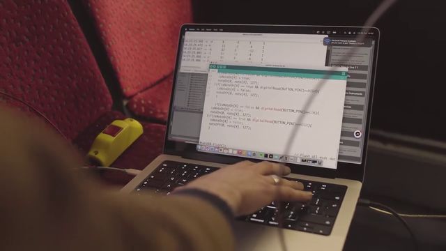 A hand typing on a laptop that shows code, set on a red bus seat beside a yellow stop button