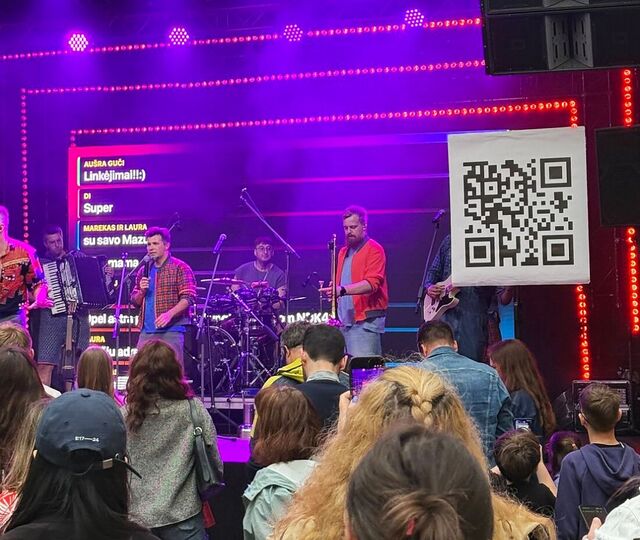 A band on stage in front of an LED screen that shows messages from the audience; the QR code hangs beside them